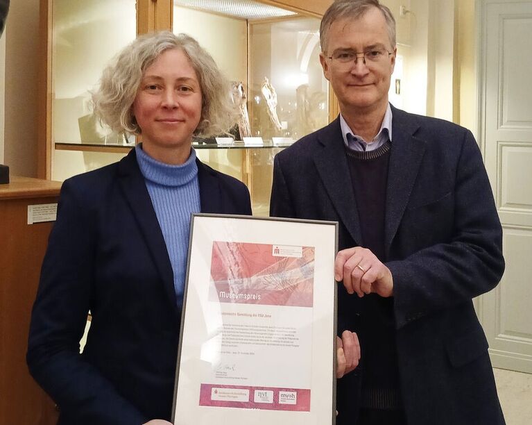 Ulrike Lötzsch und Christoph Redies zeigen die Urkunde des Museumspreises 2024. (Foto: Inka Rodigast / UKJ)